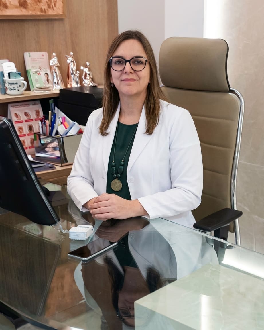 Dra. Joseane Aparecida de Brito, ginecologista em Curitiba, meia idade, cabelo channel liso e loiro médio, muito simpática olhando e sorrindo para a câmera, sentada em sua poltrona aparentemente em seu consultório. ela está de óculos e vestindo uma camiseta preta com um jaleco branco por cima, por trás uma parede em mármore bege.
