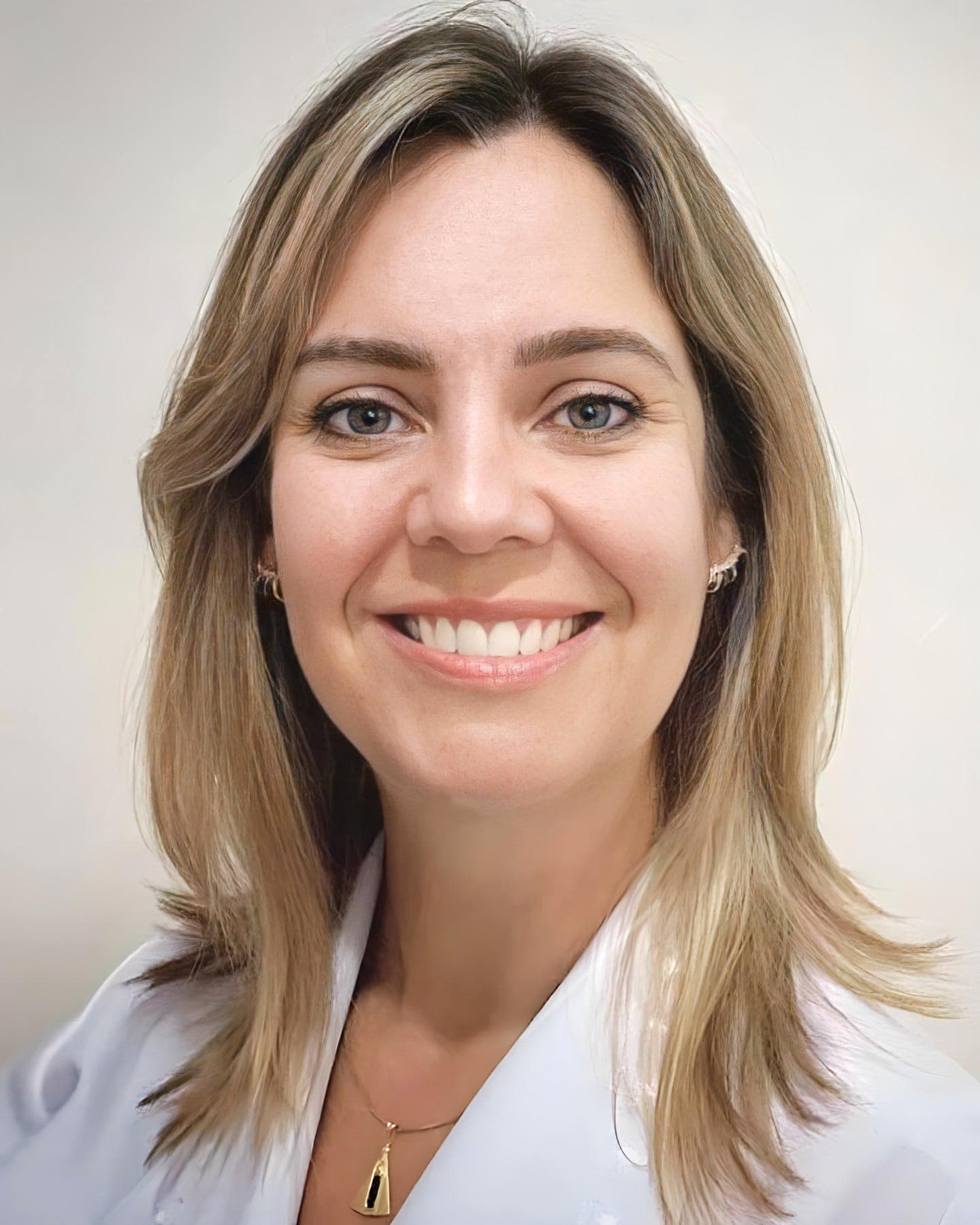 Dra. Luciana Gabardo Stahlke, médica otorrino em Curitiba, jovem, cabelos lisos e loiros até o ombro, muito simpática, sorrindo e olhando para a câmera, ela está vestindo um jaleco branco e usando um pingente, por trás uma parede lisa marrom claro.