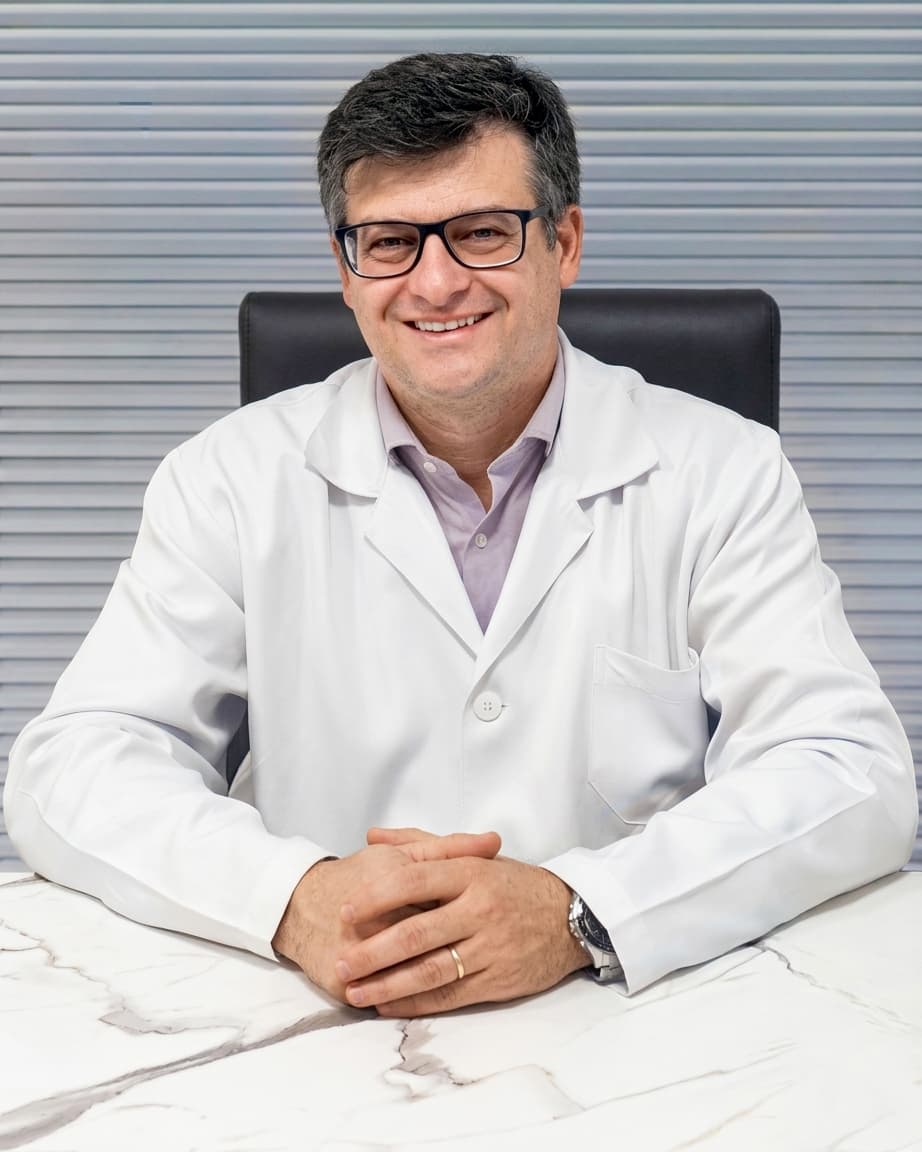Dr. Rodrigo Lazzari, cardiologista em Curitiba, meia idade, cabelos curtos e castanhos, sorrindo para a câmera enquanto está sentado, com os braços sobre sua mesa de trabalho no consultório, com as mãos juntas, ele está usando óculos, relógio e está vestindo uma camisa lilás claro com um jaleco branco por cima, por trás uma veneziana cinza fechada.