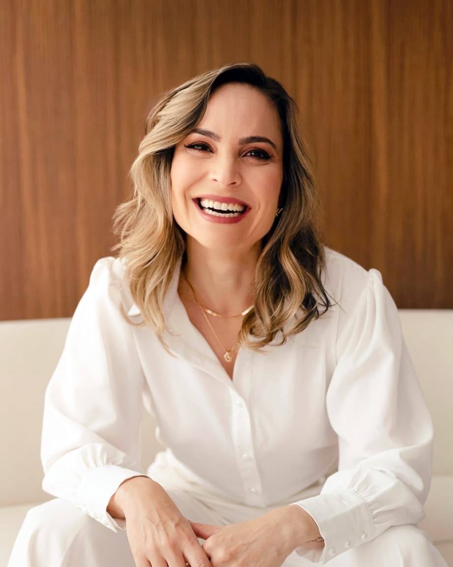 
Dra. Franciele Canesin aparece radiante em seu consultório, transmitindo profissionalismo e simpatia. Com cabelos longos e lisos de tom castanho-claro, ela sorri calorosamente para a câmera, destacando sua maquiagem impecável e batom vermelho vibrante. Vestida com um jaleco branco sobre uma camisa cinza, ela está sentada à mesa de vidro verde, com as mãos elegantemente sobrepostas. O ambiente ao fundo complementa a composição, uma parede verde, um balcão branco com uma escultura prateada e um vaso de flores que trazem um toque acolhedor. Sobre a mesa, itens que refletem sua dedicação profissional um notebook, receituário, carimbo médico e caneta.