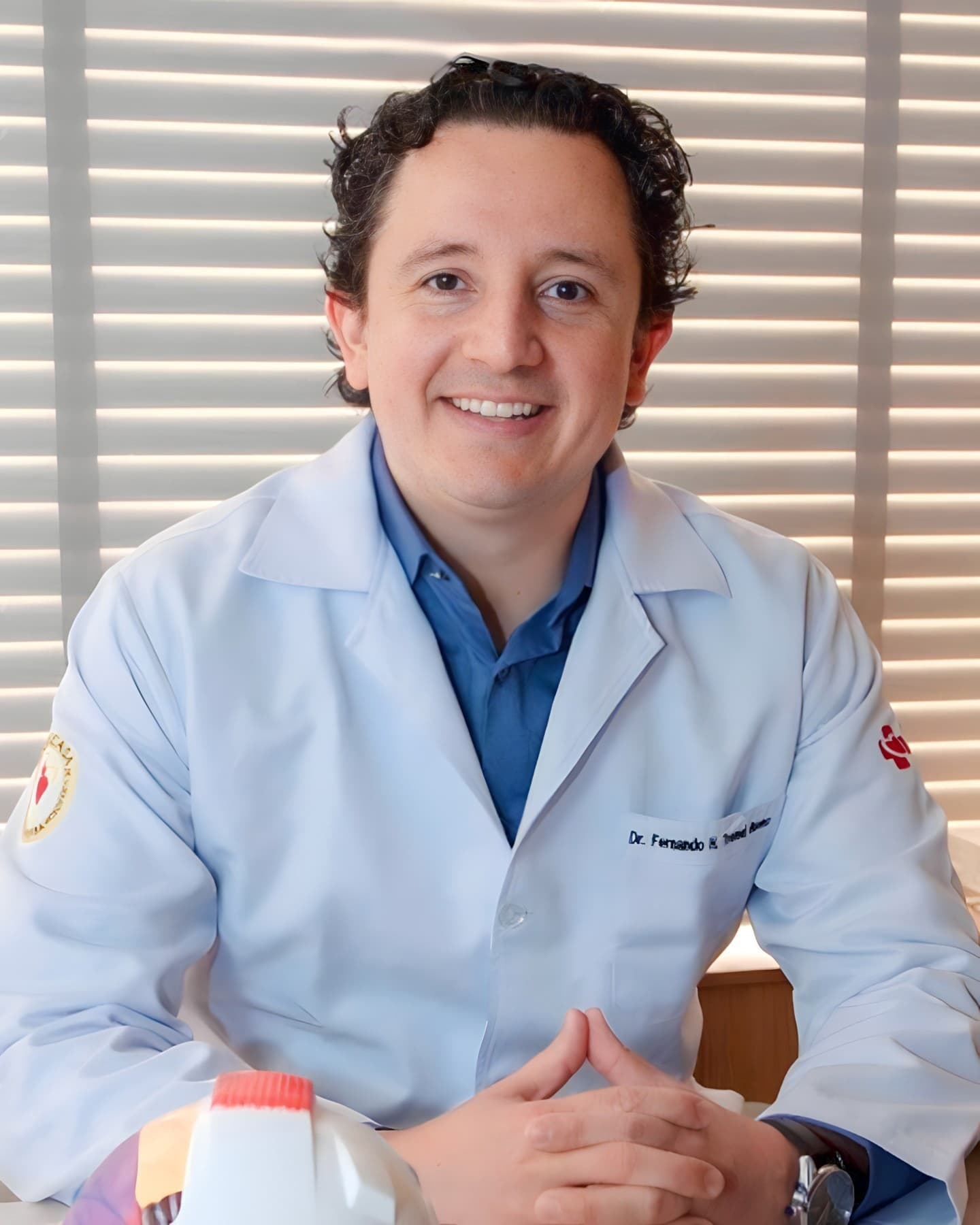 Dr. Fernando Henrique Tremel Bueno, oftalmologista, meia idade, cabelos curtos castanhos e levemte encaracolados, muito simpático sorrindo e olhando para a câmera, ele está sentado e com as mãos juntas e apoiadas sobre a mesa, aparentemente em seu consultório, ele está vestindo camisa azul com jaleco branco por cima, por trás uma janela com veneziana branca.