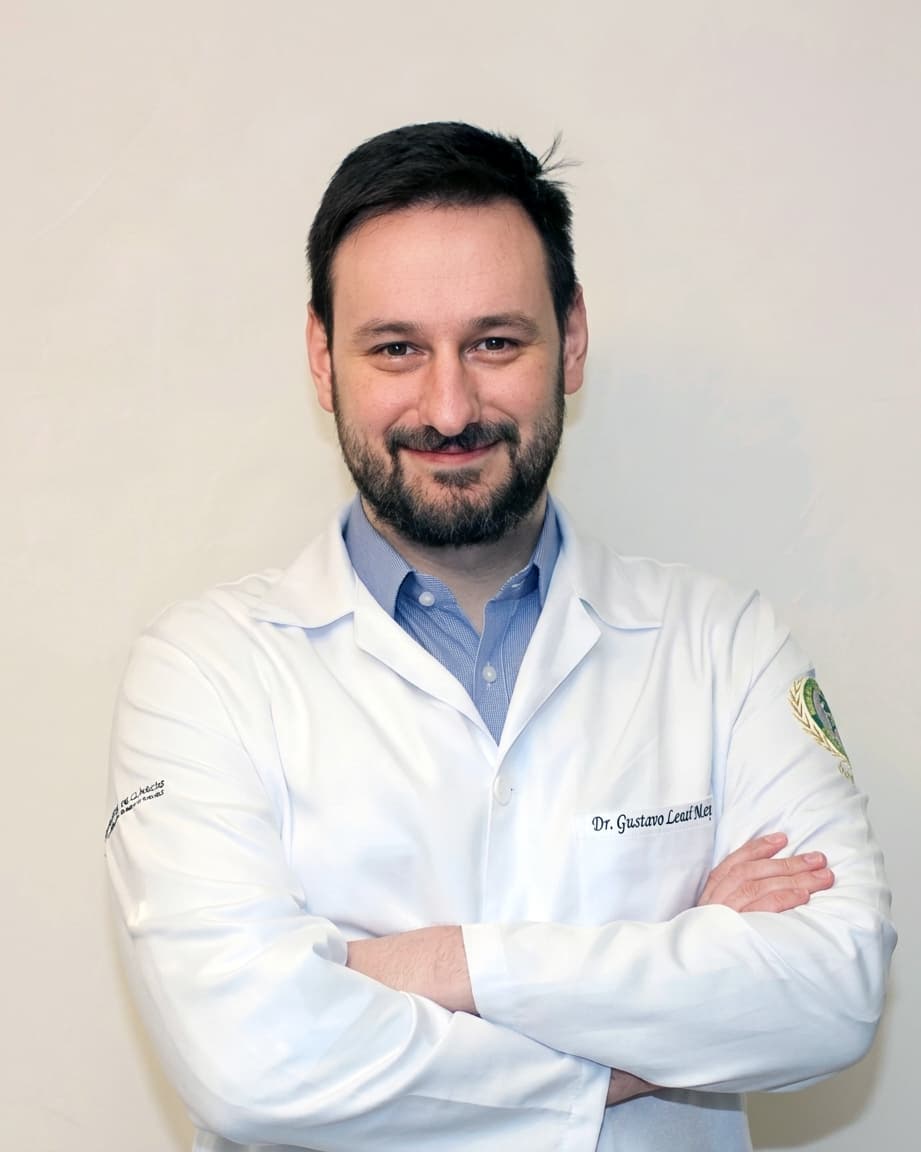 Dr. Gustavo Lenci Marques de braços cruzados, com um jaleco branco sobre uma camisa listrada azul e sorrindo para a câmera.